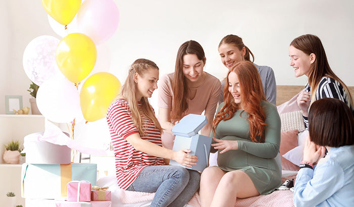 Group of Women Sitting on Bed, One With Gift Box Labeled Gift Card for Mom, Amidst Baby Shower Gifts