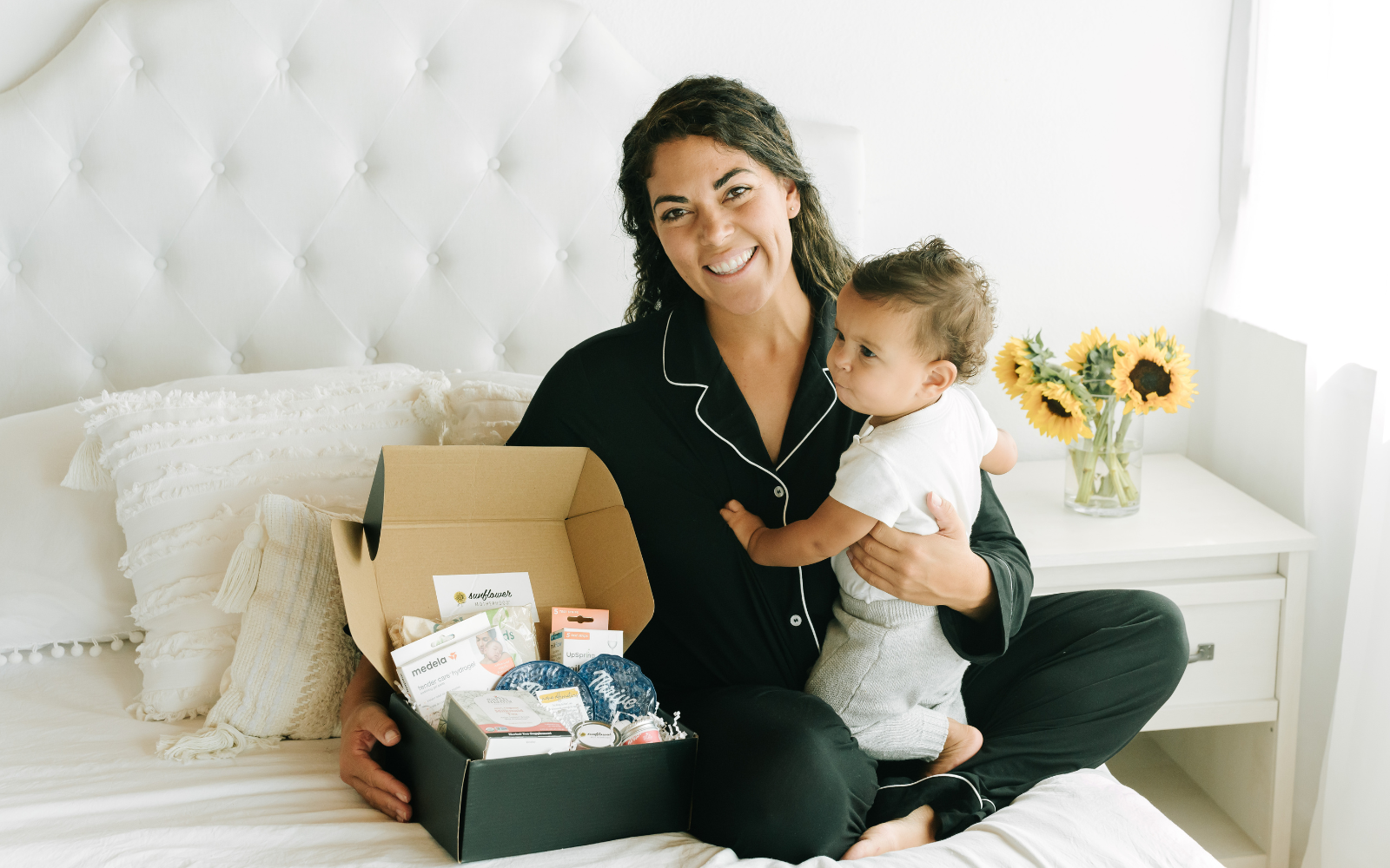 A woman holding a baby on a bed, surrounded by a box of baby products.