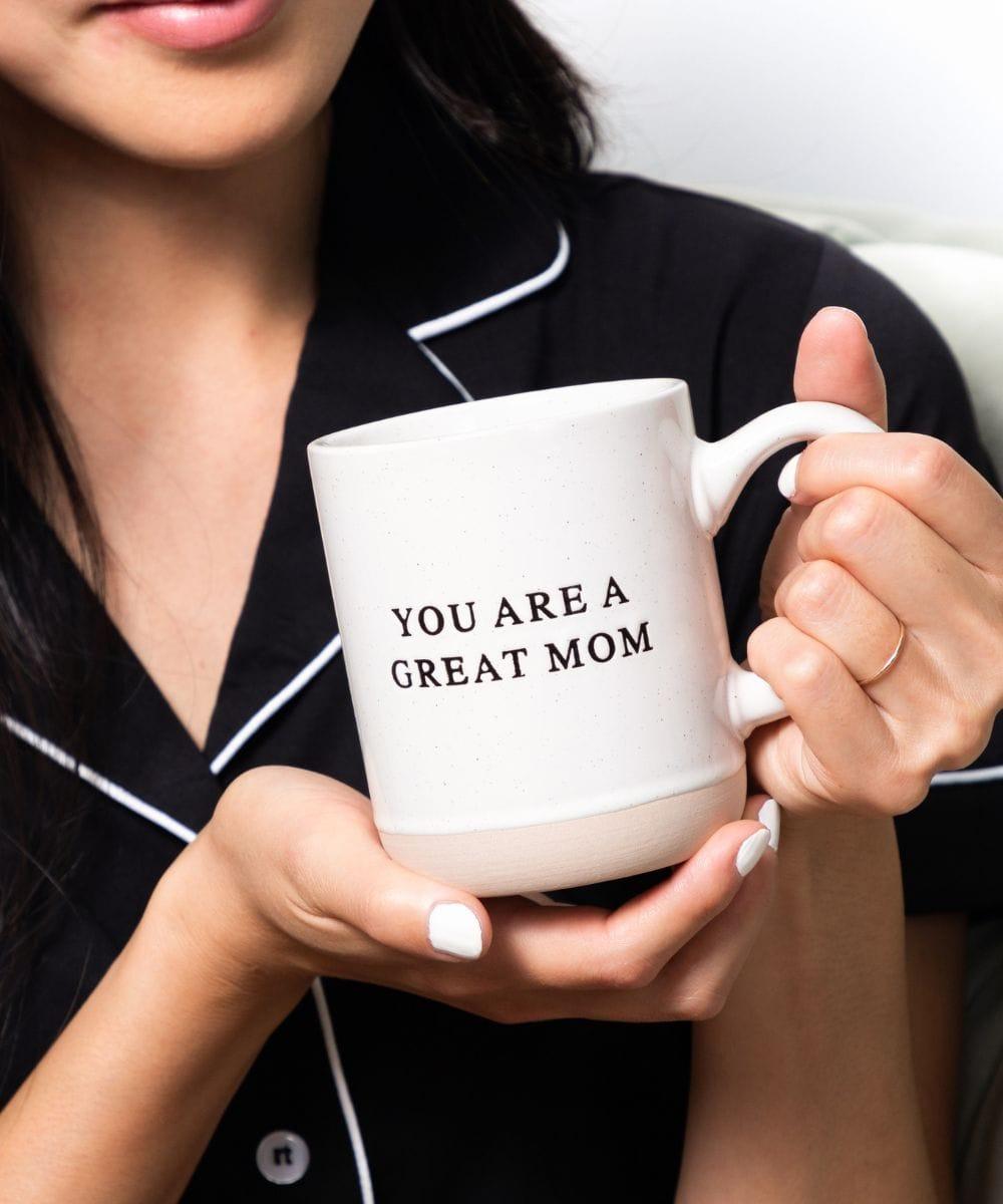 Woman smiling while holding a mug that says "you are a great mom" with a heart design, showcasing appreciation and love.