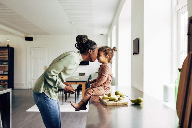 A woman kisses a child on the forehead in a modern kitchen with pears on the counter.