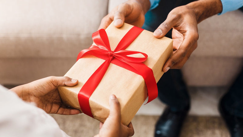 Hands exchanging a gift box wrapped in brown paper with a red ribbon bow.