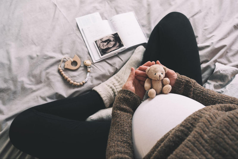 Expectant mother holding a teddy bear over her baby bump with an ultrasound image, pregnancy journal, and baby keepsake on the bed.