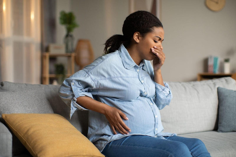 Pregnant woman in a blue shirt sitting on a couch covering her mouth with discomfort
