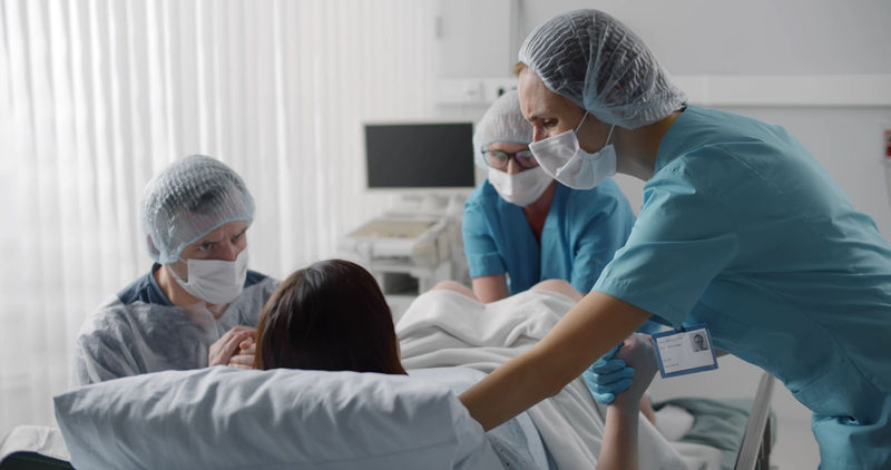 Woman in labor on hospital bed, supported by partner holding her hand. Two masked medical staff in scrubs assist during childbirth delivery.