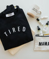 Black sweatshirt with 'tired', mug, perfume bottle, and bag on a table.