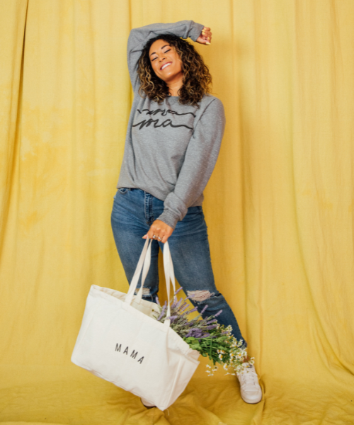 A woman in jeans and a grey sweatshirt holding a white tote bag.